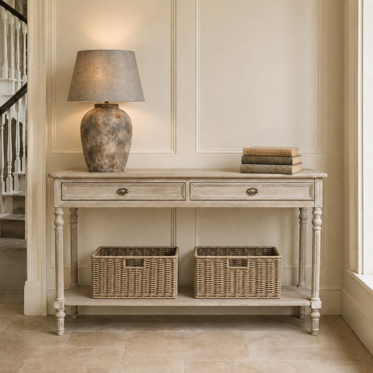 A distressed light gray wood console table with two drawers and an open lower shelf, styled with a textured ceramic table lamp and a stack of books on top, and two woven storage baskets underneath, set against a cream paneled wall.