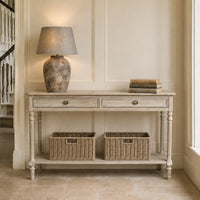 A distressed light gray wood console table with two drawers and an open lower shelf, styled with a textured ceramic table lamp and a stack of books on top, and two woven storage baskets underneath, set against a cream paneled wall.