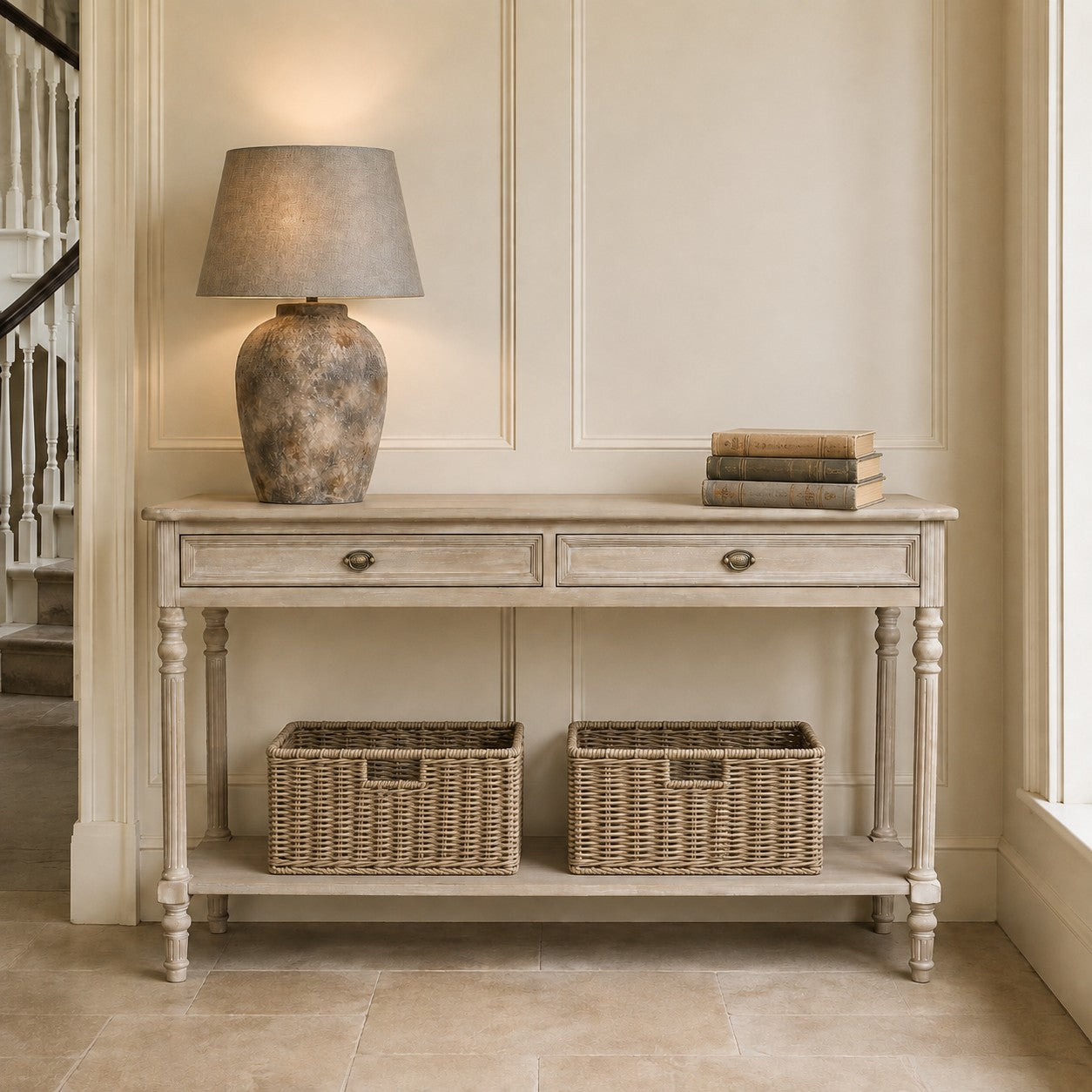 A distressed light gray wood console table with two drawers and an open lower shelf, styled with a textured ceramic table lamp and a stack of books on top, and two woven storage baskets underneath, set against a cream paneled wall.