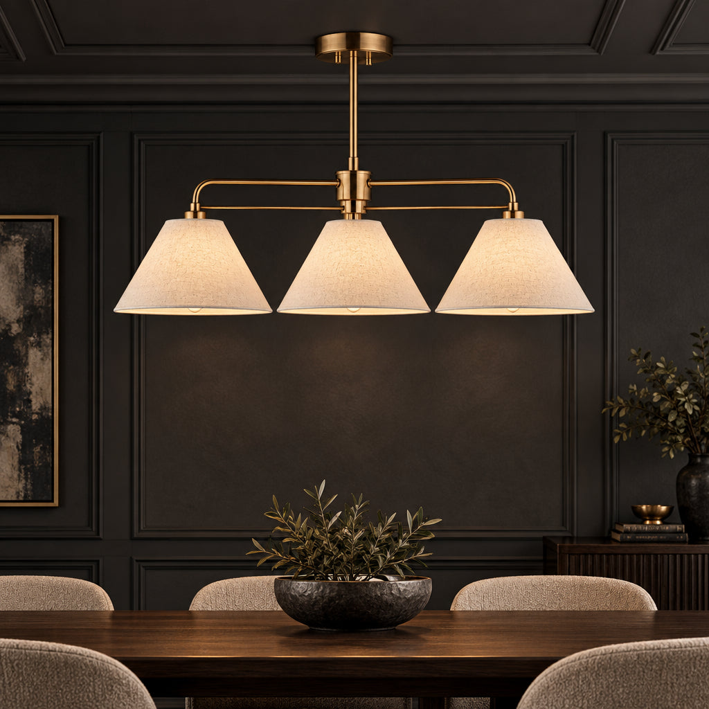 A three-light linear chandelier with warm brass-finished arms and beige fabric shades, hanging over a dark wood dining table.