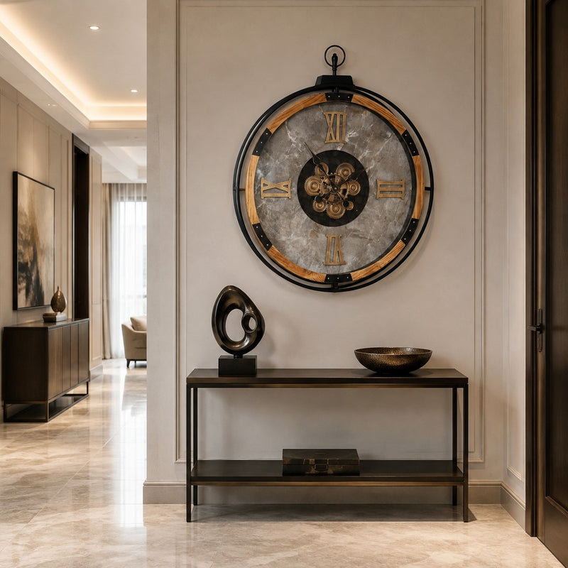 A large round wall clock with a gray stone-like face, black rim, and gold Roman numerals is mounted above a dark console table in a modern hallway with beige walls and marble flooring.