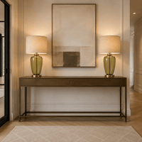 A modern console table in a warm wood finish with a black metal frame, styled in a hallway with two matching table lamps and a large abstract painting on the wall.