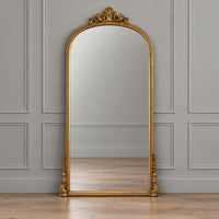 An ornate gold-framed floor mirror with an arched top and decorative scrollwork, standing against a gray paneled wall on a light wood floor.