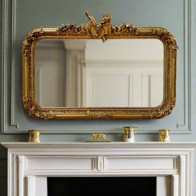 Luxurious ornate gold overmantle mirror with carved floral crest and gilt frame. Perfect statement mirror for fireplaces, hallways and elegant period interiors. In a classic living room.