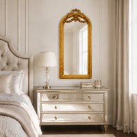 An ornate gold-framed arched mirror hanging above a mirrored dresser in a softly lit bedroom with beige bedding, a table lamp, and neutral curtains.