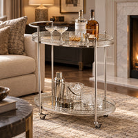 A two-tier silver metal bar cart with ornate cutout detailing and caster wheels, styled in a living room with whiskey bottles and glasses.