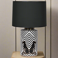 A black and white geometric pattern ceramic lamp base with black panthers, topped with a large black fabric shade, placed on a wooden surface.