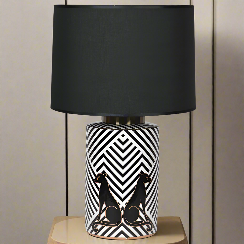 A black and white geometric pattern ceramic lamp base with black panthers, topped with a large black fabric shade, placed on a wooden surface.