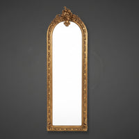 Tall, slim ornate mirror with heavy gilt frame on a neutral background.