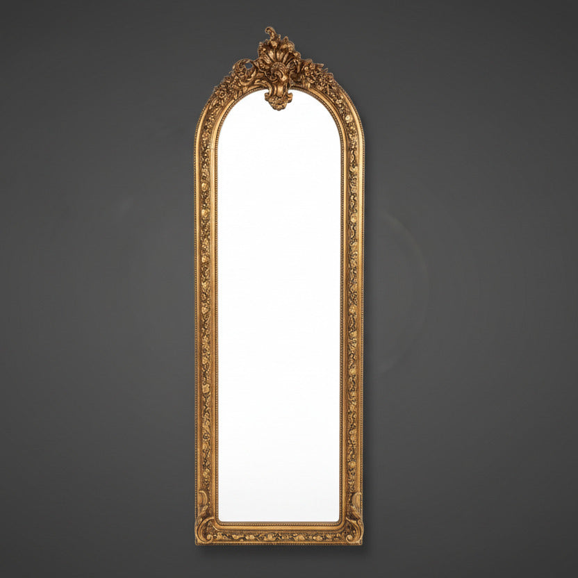 Tall, slim ornate mirror with heavy gilt frame on a neutral background.