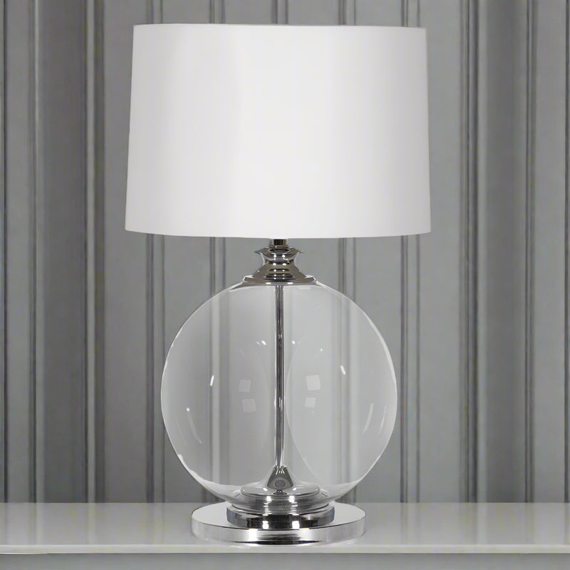 An elegant, round glass based table lamp with a white shade. The clear glass set into a nickel metal base and neck.