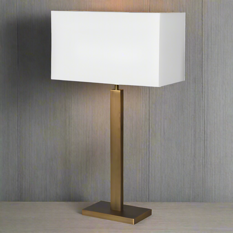 A tall gilt metal lamp with a rectangular white shade with a gold lining on a wooden surface.