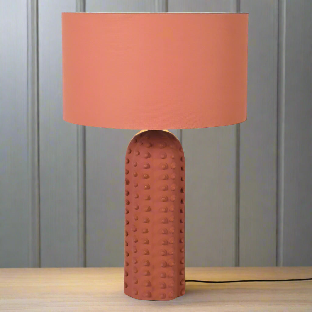 This is a ceramic table lamp finished in a warm terracotta color, featuring a monochromatic design where both the base and the drum-shaped fabric shade share the same earthy hue. The lamp base stands out with its raised bobble texture, giving it a tactile and contemporary appeal. The base is tall and cylindrical with a rounded top, and the bobbled surface adds visual depth and a handcrafted feel, while the uniform terracotta finish provides a cohesive, minimalist look. This lamp is ideal for interiors with 