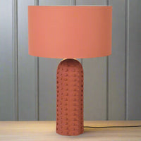 This is a ceramic table lamp finished in a warm terracotta color, featuring a monochromatic design where both the base and the drum-shaped fabric shade share the same earthy hue. The lamp base stands out with its raised bobble texture, giving it a tactile and contemporary appeal. The base is tall and cylindrical with a rounded top, and the bobbled surface adds visual depth and a handcrafted feel, while the uniform terracotta finish provides a cohesive, minimalist look. This lamp is ideal for interiors with 