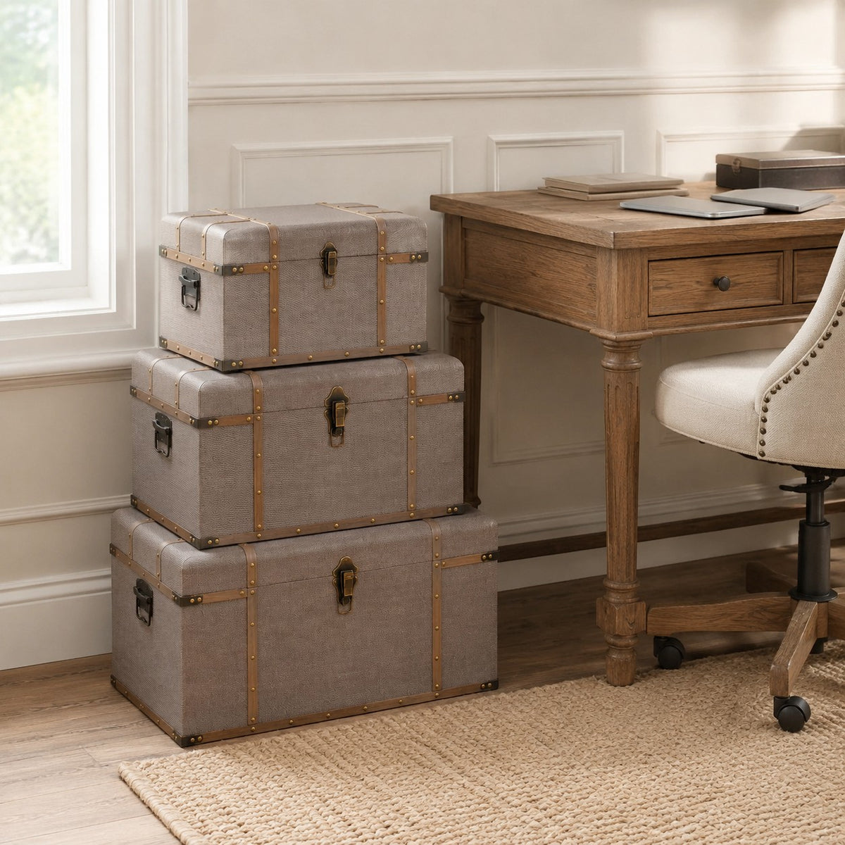Three stacked gray fabric storage trunks with brass hardware in a home office setting next to a wooden desk and chair.