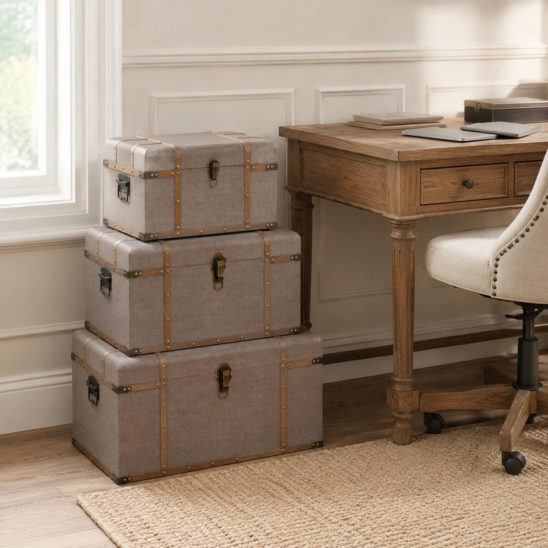 Three stacked gray fabric storage trunks with brass hardware in a home office setting next to a wooden desk and chair.