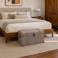 A gray upholstered storage trunk with brass hardware sits at the foot of a wooden bed in a styled bedroom setting.