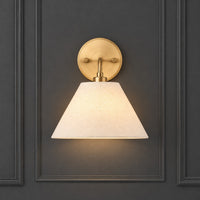 A wall-mounted sconce with a warm brass circular backplate and a tapered off-white fabric shade, shown lit against a dark gray paneled wall.