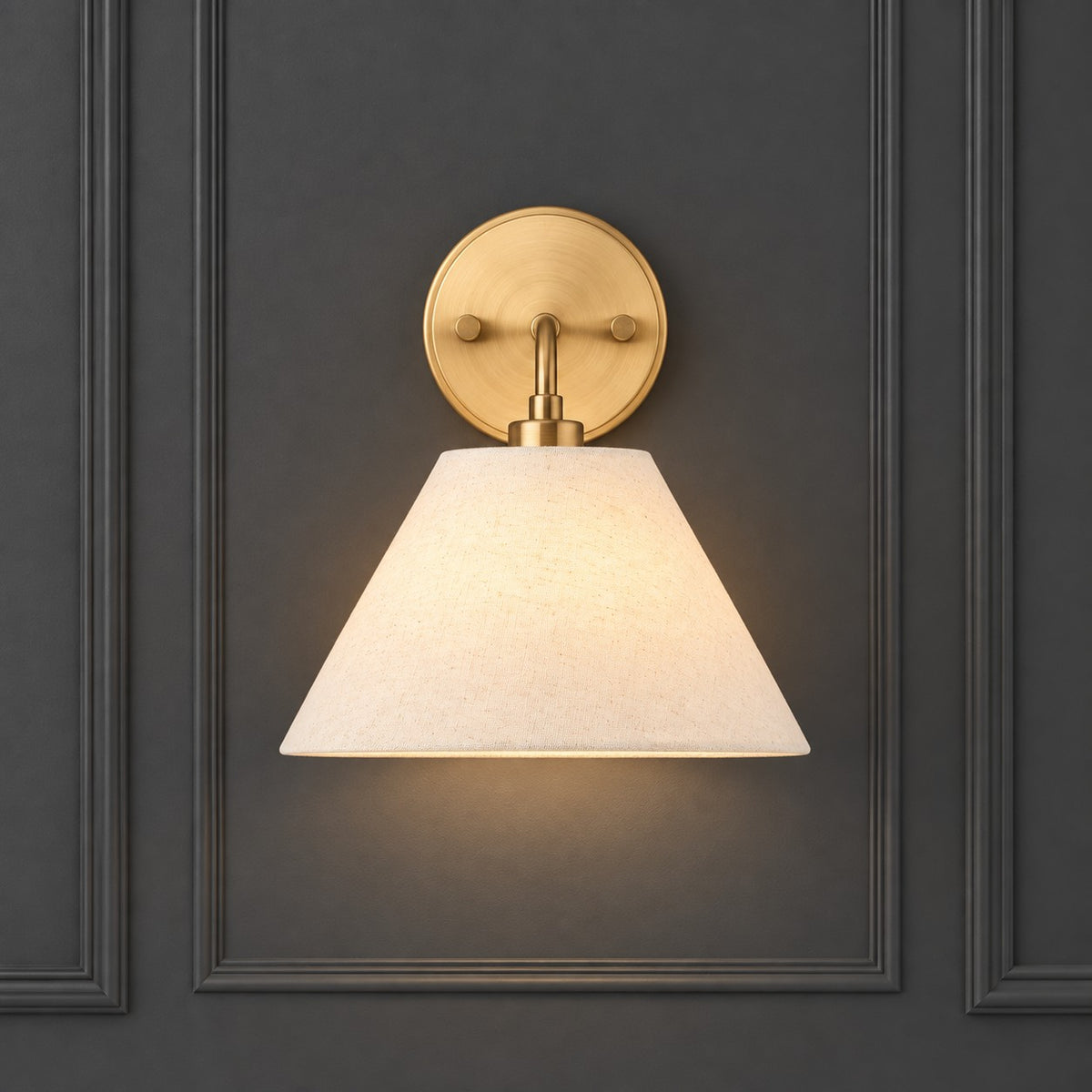 A wall-mounted sconce with a warm brass circular backplate and a tapered off-white fabric shade, shown lit against a dark gray paneled wall.