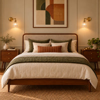 A styled bedroom scene featuring a wooden bed frame with a upholstered headboard, dressed in cream bedding, olive green pillows, and a green throw blanket. The bed is flanked by matching wooden nightstands with lit wall sconces.