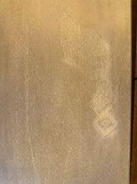 A close-up of a light beige or cream-colored textured surface with subtle vertical grain and a faint smudge or stain near the lower right area.