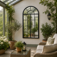Arched black, open-door style window mirror in a conservatory with outdoor furniture. 